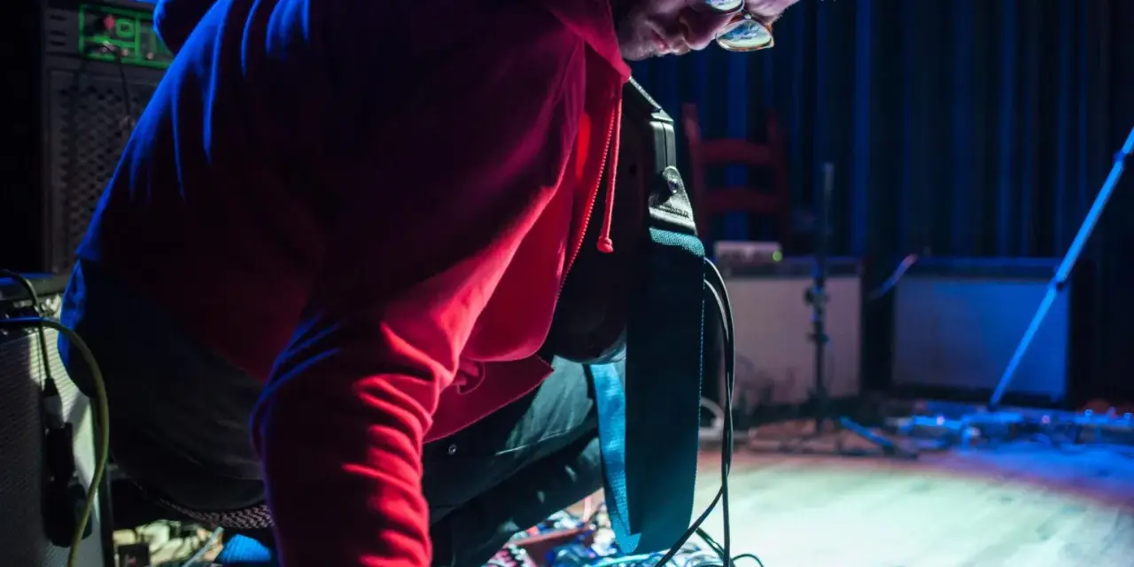 Musiker mit roten Kapuzenpullover beugt sich über ein Pedalboard, um den "Delay"-Effekt einzustellen.