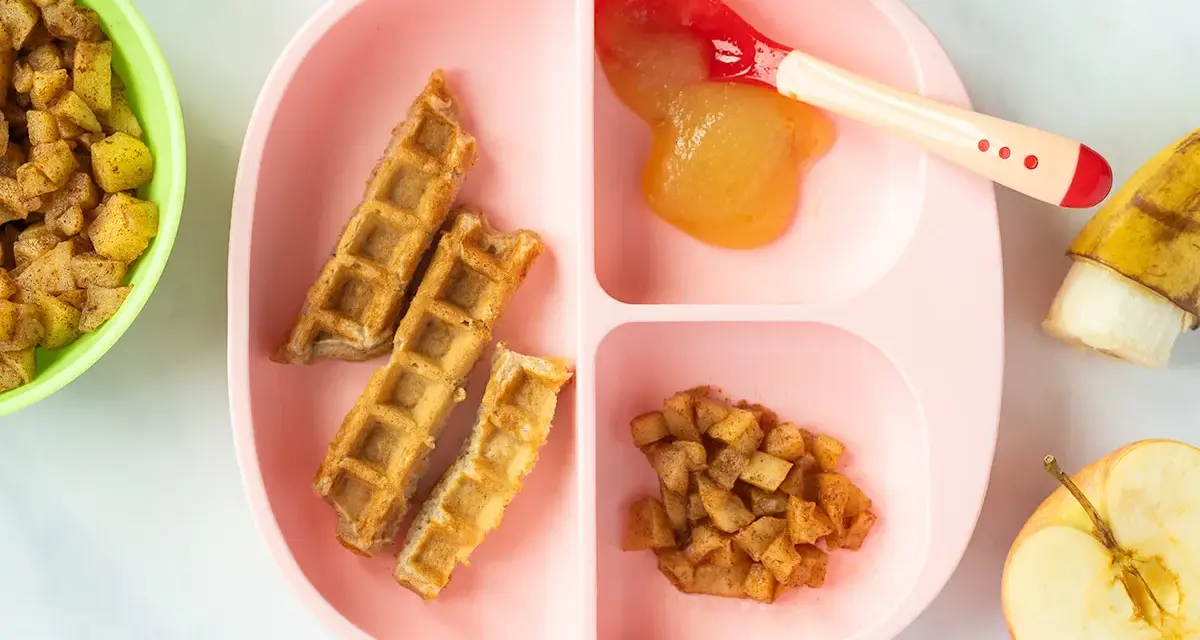 Baby-freundliche Apfelwaffeln mit Apfelmus und Zimt-Apfelstückchen auf einem rosa Teller. Ein halber Apfel und eine Banane liegen daneben.