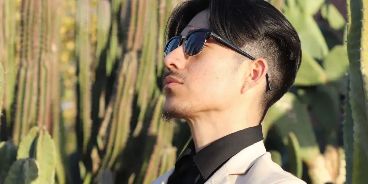 Uomo con abito beige e camicia nera, occhiali da sole, tra i cactus. Un look elegante per ogni occasione.
