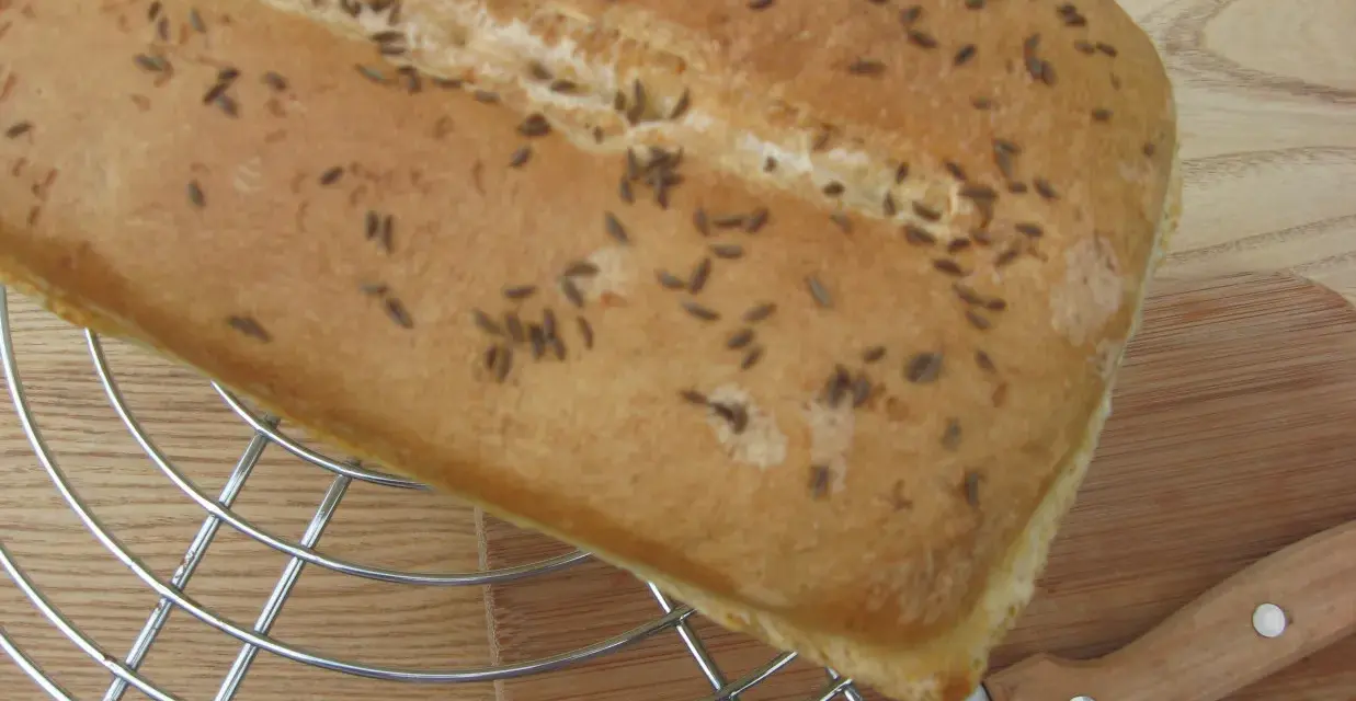 Chleb z kminkiem na metalowej kratce, obok nóż i deska. Pachnący, świeży wypiek gotowy do krojenia.