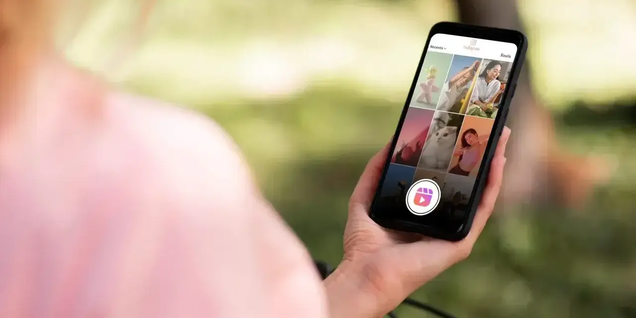 Osoba trzyma telefon z widokiem na ekran Instagrama, gdzie widać siatkę zdjęć i ikonę Reels. Dowiedz się, jak zrobić rolkę na Instagramie ze zdjęć.