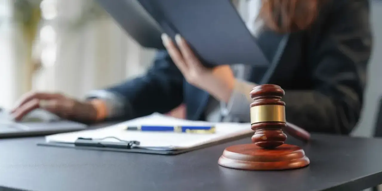 Sądowy młotek i dokumenty na biurku. Prawnik w tle studiuje akta, zastanawiając się, czy radca prawny może być obrońcą.