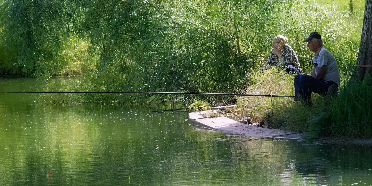 Dwaj wędkarze siedzą nad zieloną wodą, czekając na branie. Jeden z nich, starszy mężczyzna, skupiony na swoim sprzęcie, gotowy do **casting spinning**.