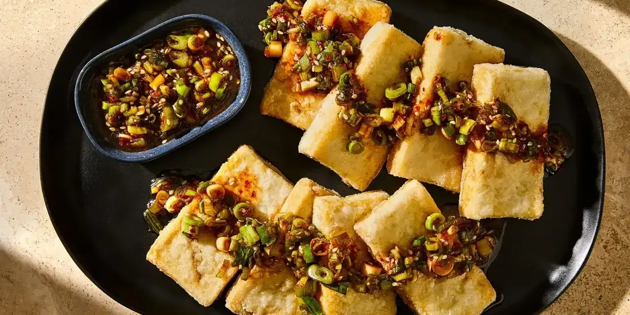 Chrupiące kawałki tofu, polane aromatycznym sosem z dymką i sezamem, podane na czarnym talerzu.