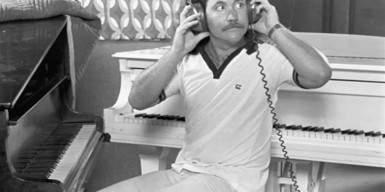 Billy Powell hört mit Kopfhörern Musik, während er neben einem Klavier sitzt.
