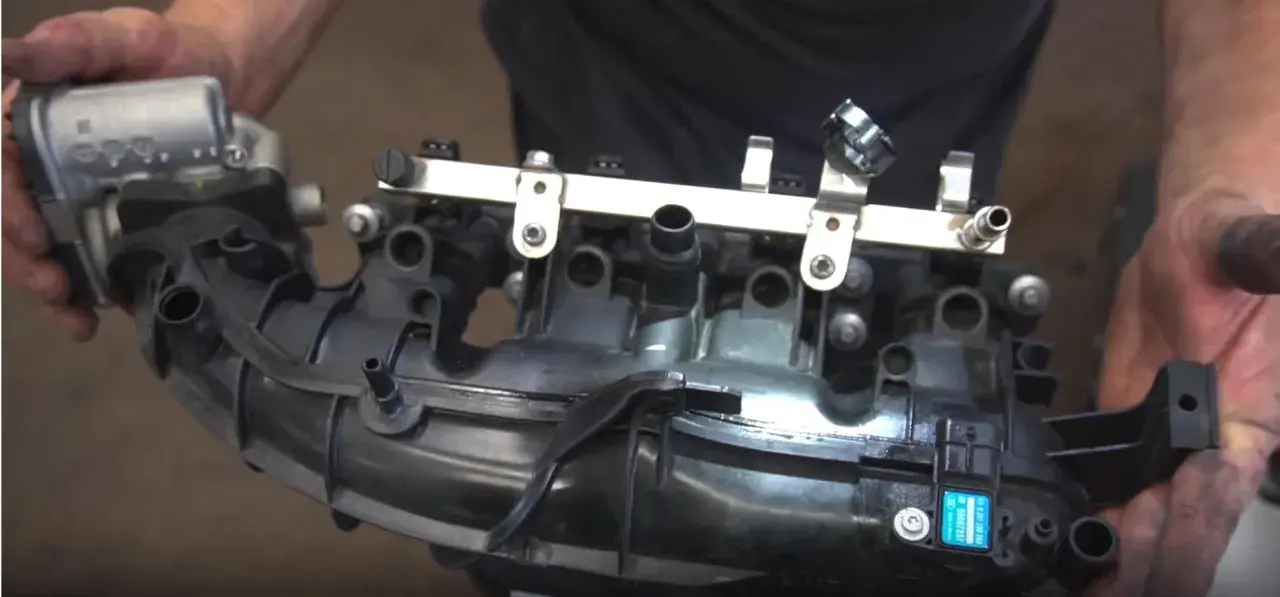 Hands hold a black plastic intake manifold with a fuel rail and sensors. This part is crucial for intake manifold runner control.