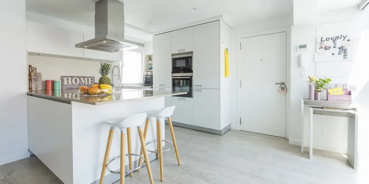 Moderna cocina blanca y gris claro con isla central, taburetes y frutas frescas.