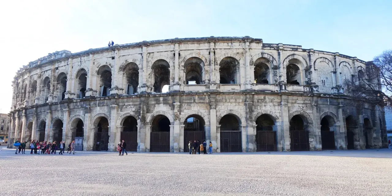 The ancient Roman amphitheater in Nîmes, France, stands majestically under a clear blue sky. Tourists gather in the plaza, ready to explore this historic marvel.