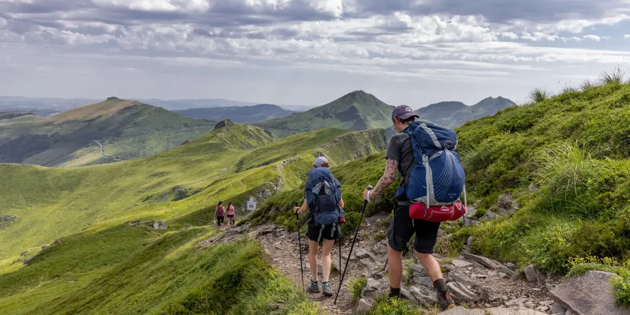 Randonnée sur le Puy Mary, des marcheurs explorent un sentier rocailleux bordé de verdure, avec des montagnes ondulantes en arrière-plan sous un ciel nuageux.