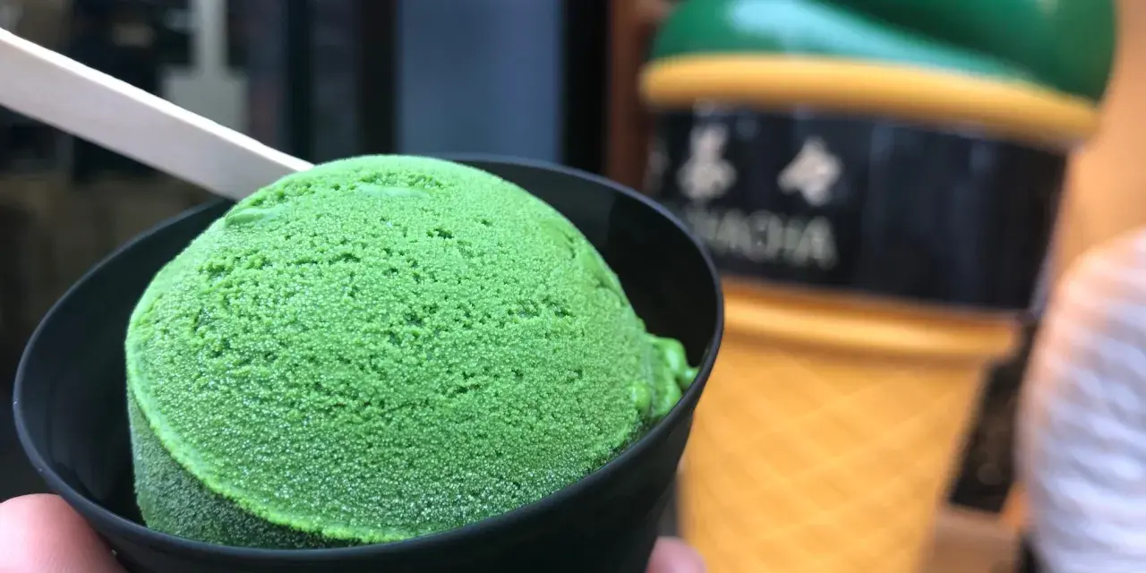Zielone, japońskie lody matcha w czarnym kubeczku, z drewnianą łyżeczką. W tle wielki, żółty rożek z lodami.