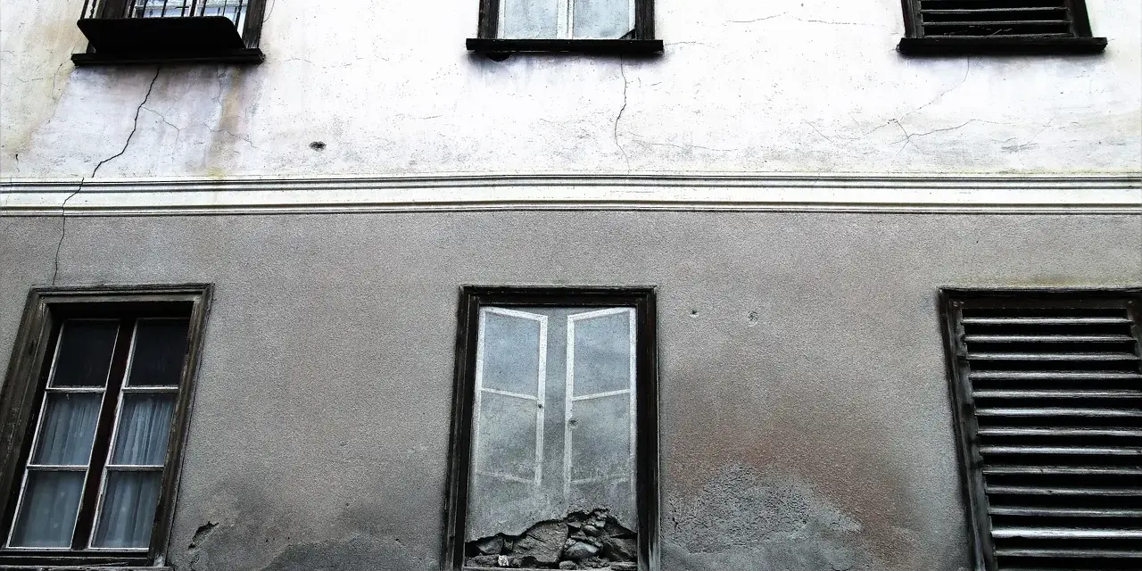 Stara elewacja z widocznymi pęknięciami i zaciekami. Okna z żaluzjami i jedno wybite.