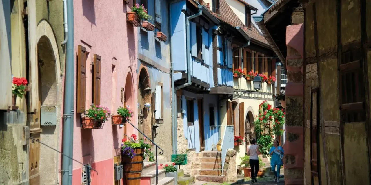 Charming street with colorful houses, perfect for day trips from Strasbourg. Two women stroll past flower-filled window boxes.