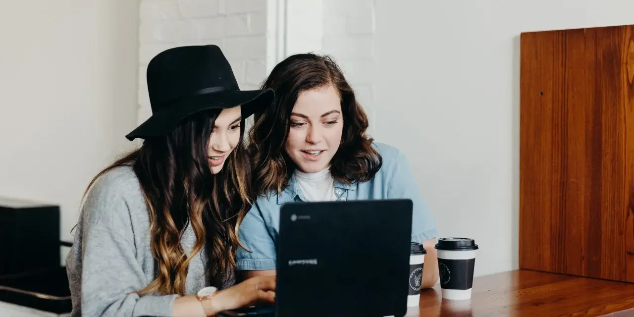 Dwie młode kobiety pracują na laptopie, jedna w kapeluszu, druga w dżinsowej koszuli. Ich wspólny projekt to passe recent.