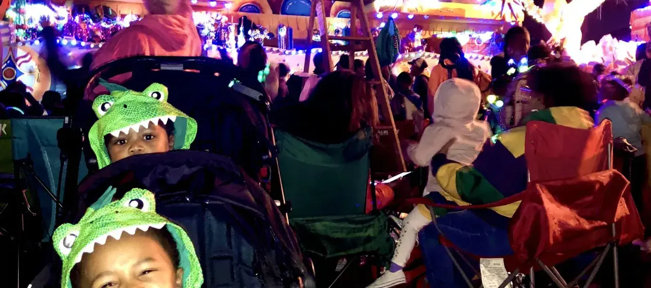 Two kids in dinosaur hoodies watch a vibrant Mardi Gras parade. They're ready for fun at this family-friendly event.