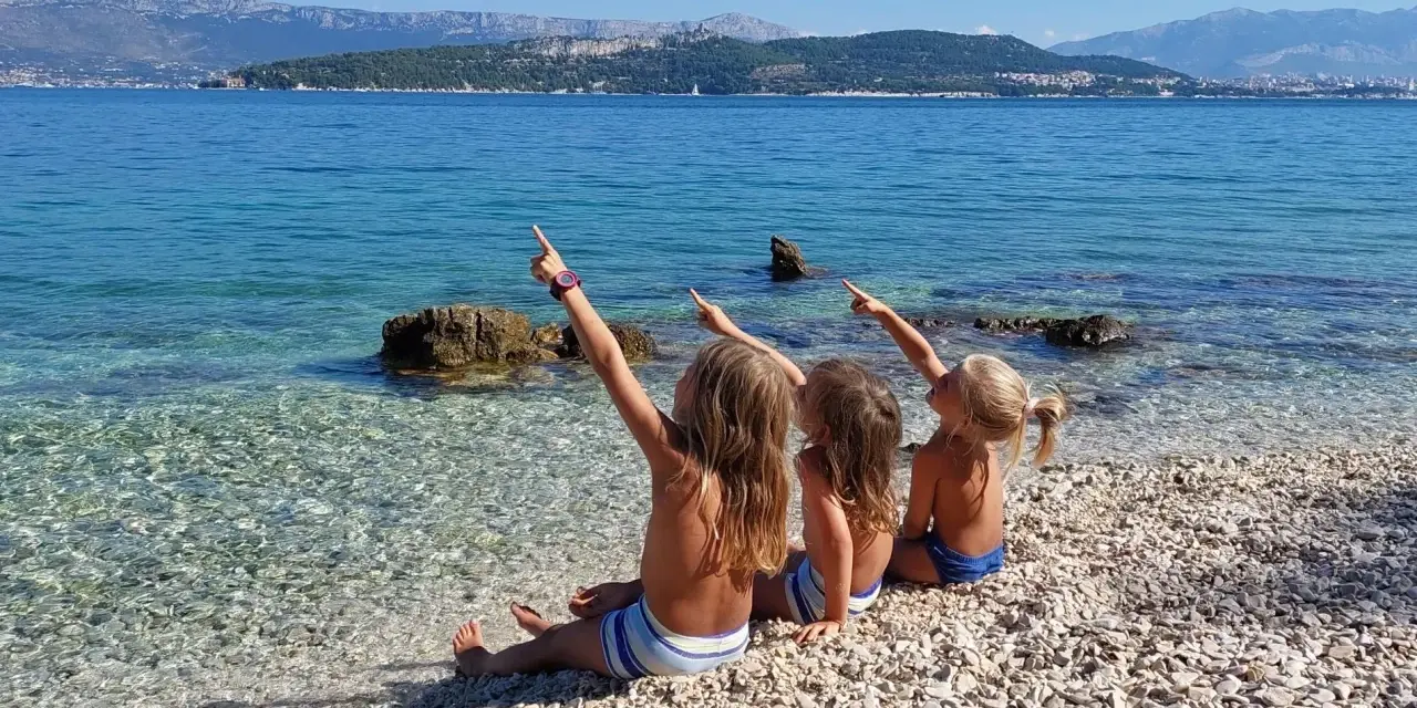 Trzy dziewczynki siedzą na kamienistej plaży w Chorwacji, wskazując palcami na morze. Idealne miejsce, gdzie do Chorwacji z dziećmi.