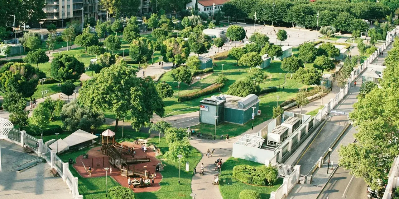 Park z placem zabaw i alejkami, otoczony budynkami. Wygląda jak jeden z najbardziej zielonych miast w Polsce.