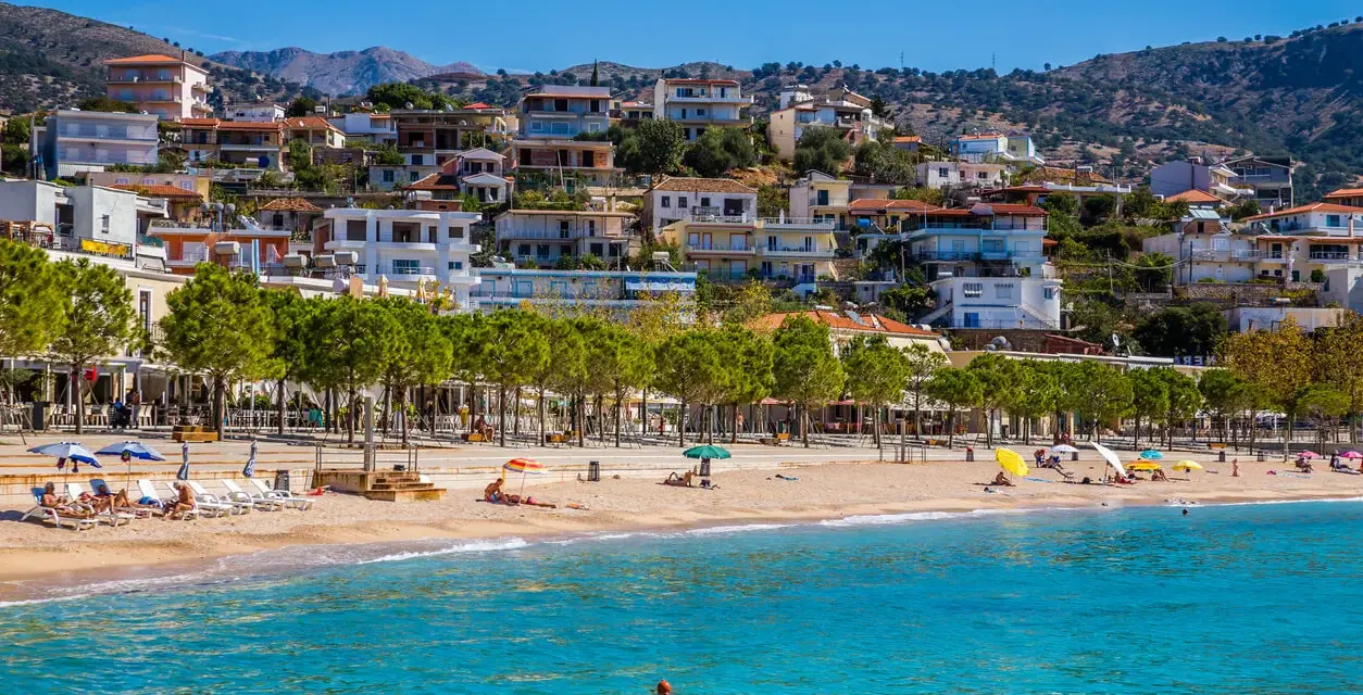 Albania, pogoda czerwiec: słoneczny dzień na plaży, turkusowe morze, piaszczysty brzeg, zielone drzewa i białe budynki na wzgórzu.