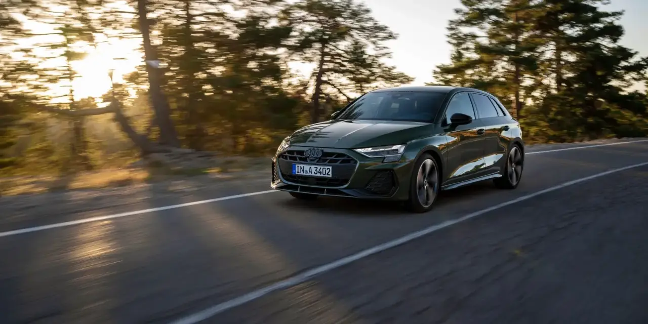 Un'auto verde scuro, un segmento premium, sfreccia su una strada panoramica al tramonto, con alberi sfocati sullo sfondo.