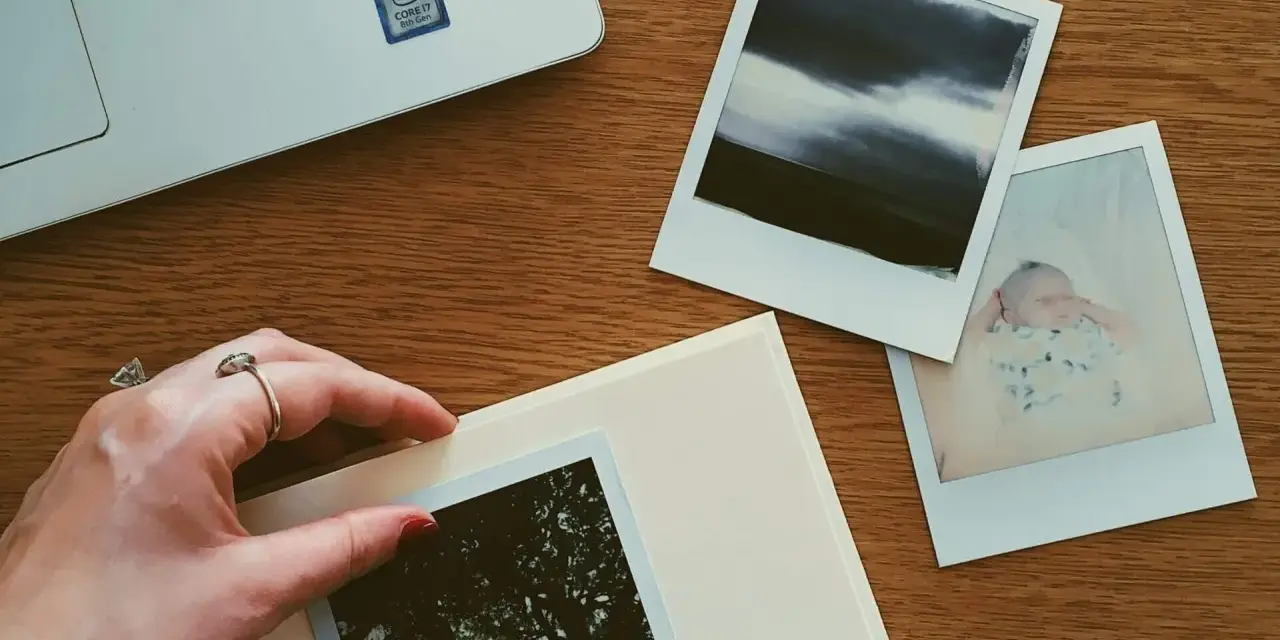 Dłoń układa zdjęcie na albumie, obok inne fotografie i laptop. Idealne miejsce, gdzie przechowywać zdjęcia z telefonu.