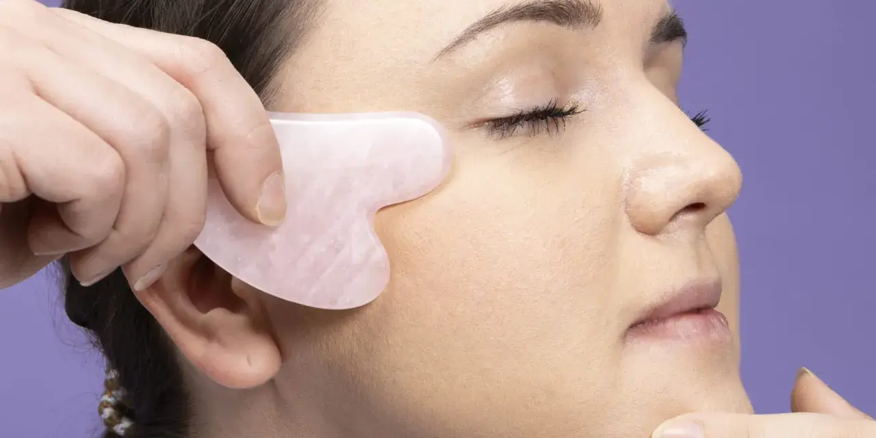 Eine Hand nutzt ein Gua Sha Stein für eine Gesichtsmassage. Das Werkzeug ist herzförmig und aus rosafarbenem Stein.