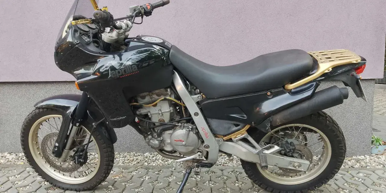 Czarny motocykl Aprilia Pegaso 650 z złotym bagażnikiem i lusterkami, zaparkowany na kostce brukowej.