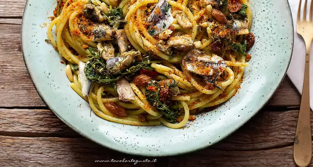 Un piatto di pasta con le sarde, condita con erbe aromatiche, pinoli e pangrattato tostato, servita su un piatto azzurro.