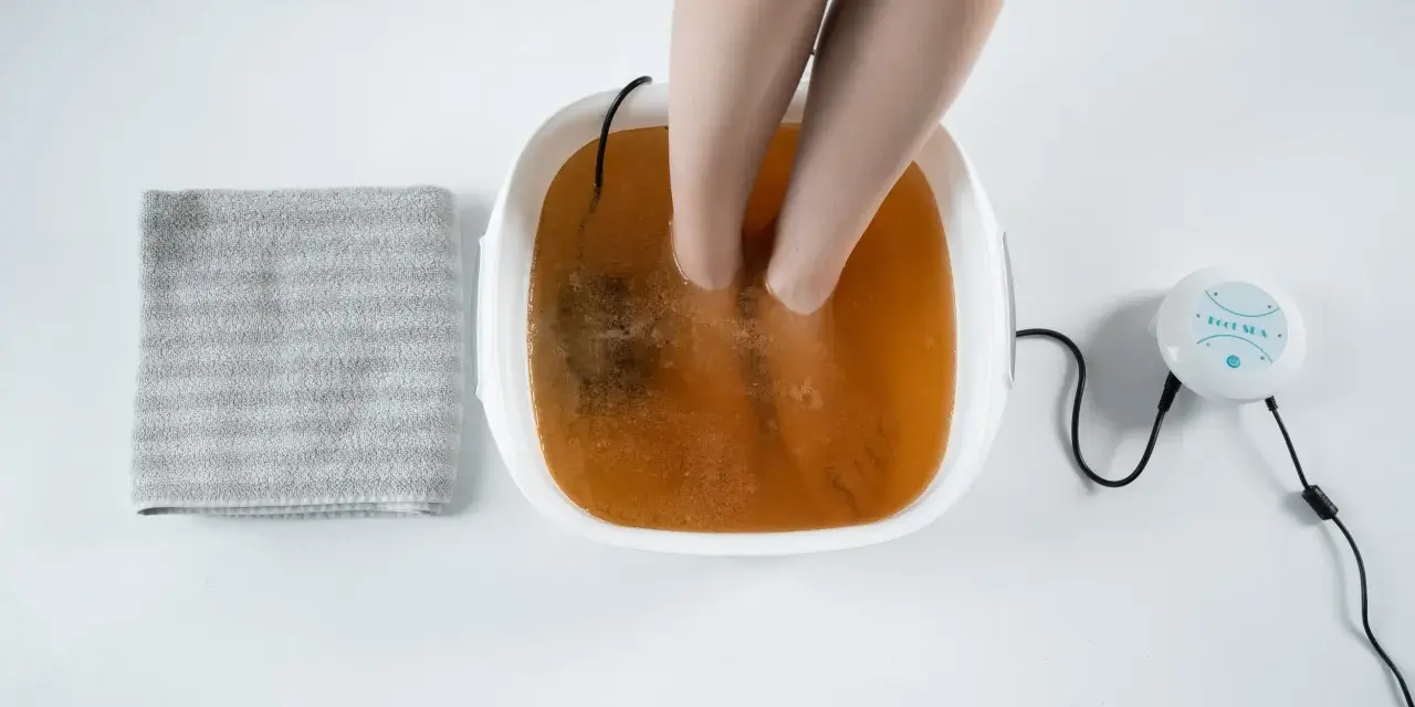 A person's feet are soaking in a foot spa with a brown solution, suggesting a method for how to remove toxins from feet naturally. A towel is nearby.