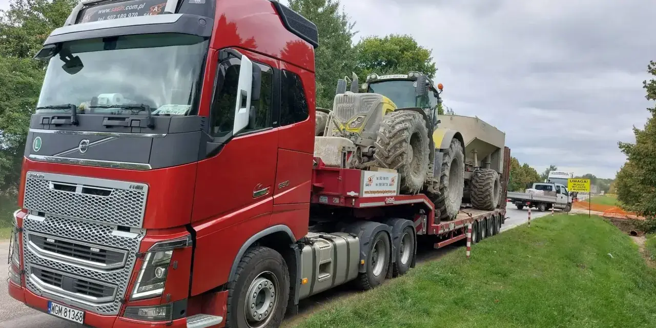 Czerwony ciągnik siodłowy przewozi koparkę na naczepie. To transport ponadgabarytowy, który wymaga specjalnych zezwoleń i przygotowania.