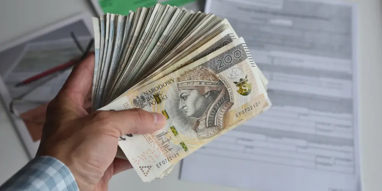 Dłoń trzyma plik banknotów 200 zł. W tle dokumenty, być może dotyczące tego, ile godzin to 0.5 etatu.