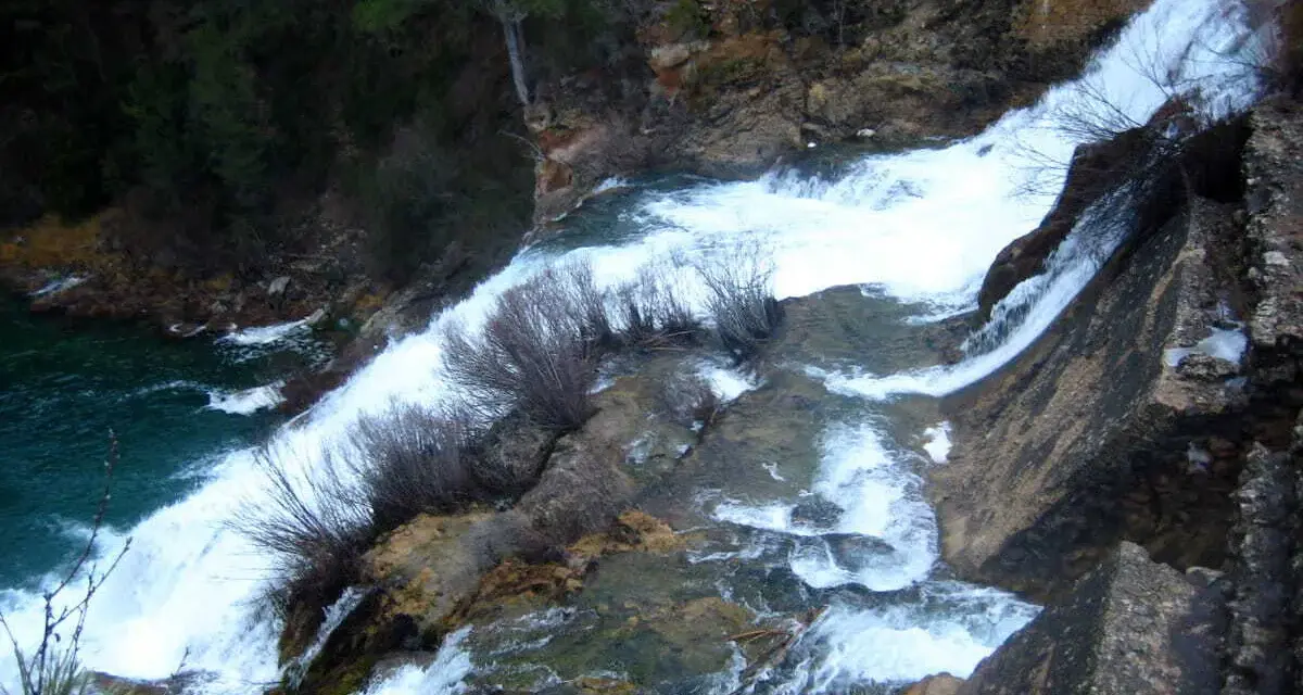 El salto de Poveda, una cascada de agua blanca y espumosa que desciende por rocas y vegetación, desemboca en un río de aguas turquesas.