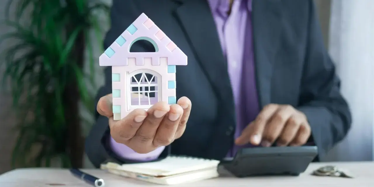 Hombre con traje calcula cuánto puede subir el alquiler, sosteniendo una casa de juguete.