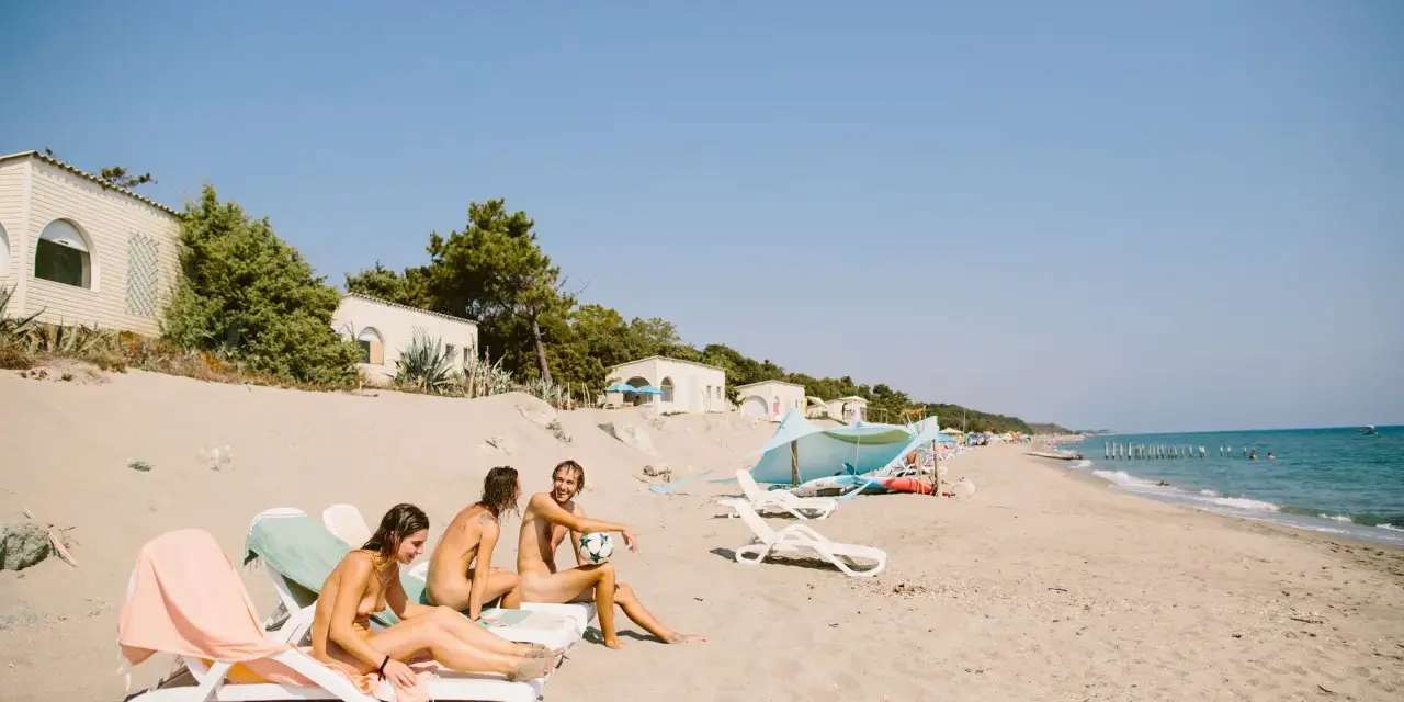 Trzy osoby relaksują się na plaży w nudystycznym obozie, siedząc na leżakach. Jeden z nich trzyma piłkę.