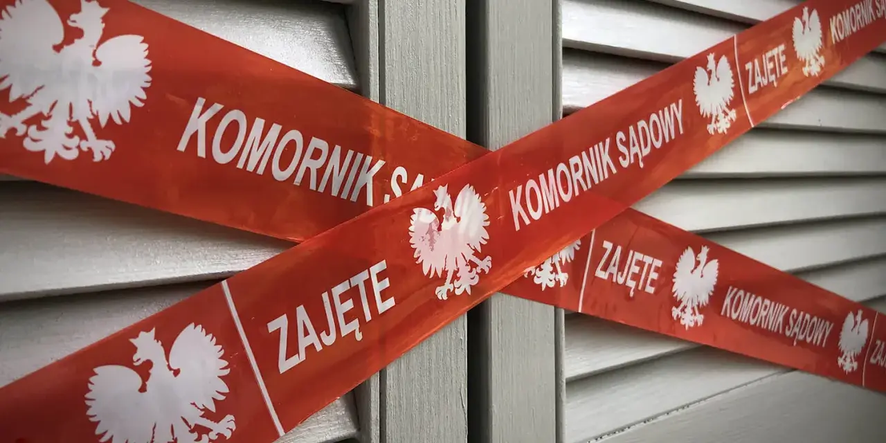 Czerwona taśma z napisem "KOMORNIK SĄDOWY" i godłem Polski krzyżuje się na szarych drzwiach. Nakaz zapłaty z sądu kiedy komornik może oznaczać zajęcie.