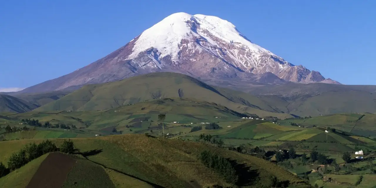 Pokryty śniegiem wulkan Chimborazo, jeden z najwyższych gór świata, góruje nad zielonymi, pofałdowanymi wzgórzami z polami uprawnymi.