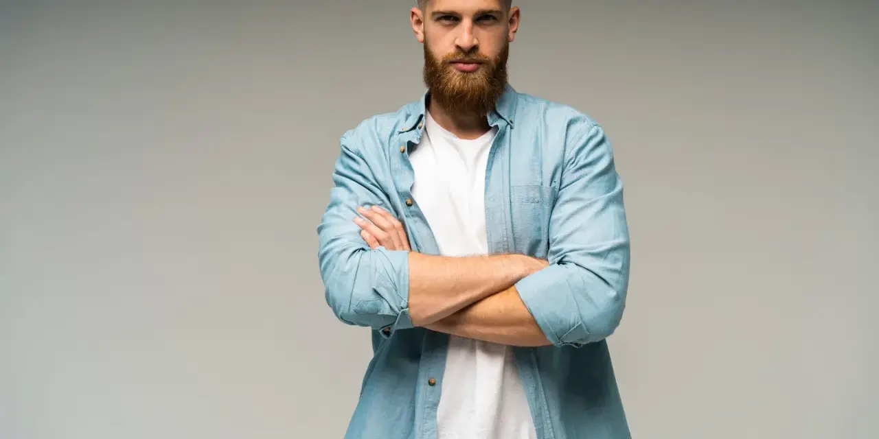 Uomo con barba e capelli biondi, camicia azzurra aperta su t-shirt bianca, braccia conserte. Look trasandato ma curato.