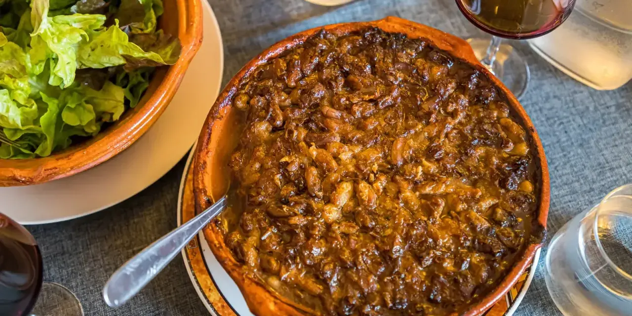 Cassoulet de Castelnaudary burbujeante en cazuela de barro, acompañado de ensalada verde y vino tinto.
