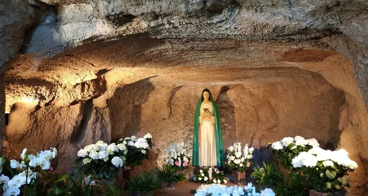 A statue of Mary stands in a grotto, surrounded by lush white flowers. The cave walls are illuminated, creating a serene atmosphere, reminiscent of the three fountains of grace.