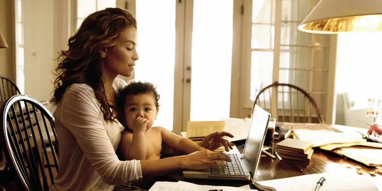 Una mamma lavoratrice con il suo bambino in grembo, mentre digita al computer tra libri e fogli.