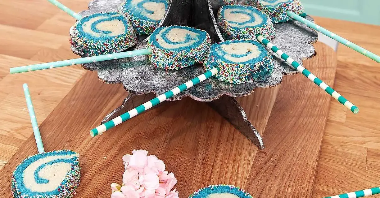 Blå och vita cake pops med strössel på ett silverfärgat fat. Vissa ligger på ett träunderlag med rosa blommor.