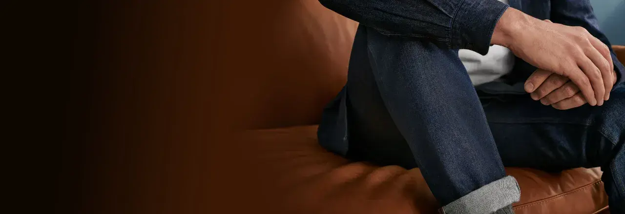 Uomo con jeans scuri seduto su divano in pelle. Scopri come lavare jeans a mano per mantenerli perfetti.