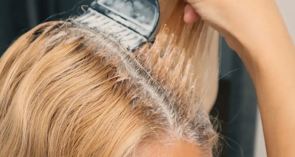 Dłoń z pędzlem nakłada rozjaśniacz na blond włosy, pokazując, jak rozjaśnić włosy w domu.