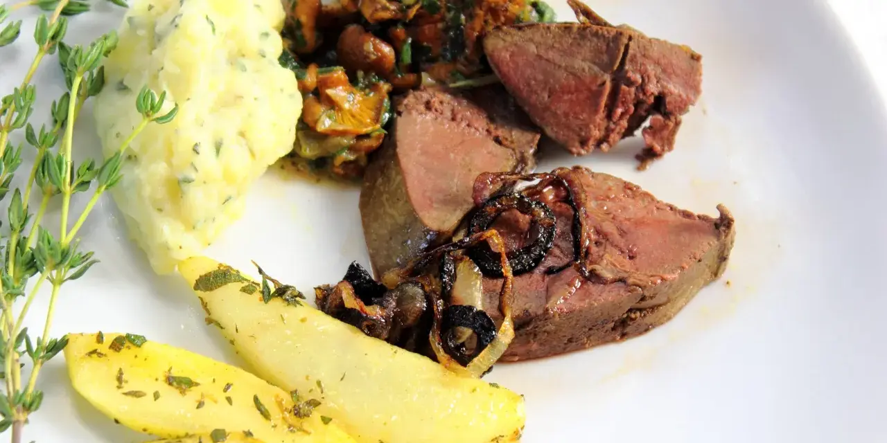 Rehleber zubereiten: zarte Scheiben Rehleber mit gebratenen Zwiebeln, Kartoffelpüree, gebratenen Kartoffelspalten und frischen Kräutern auf einem weißen Teller.