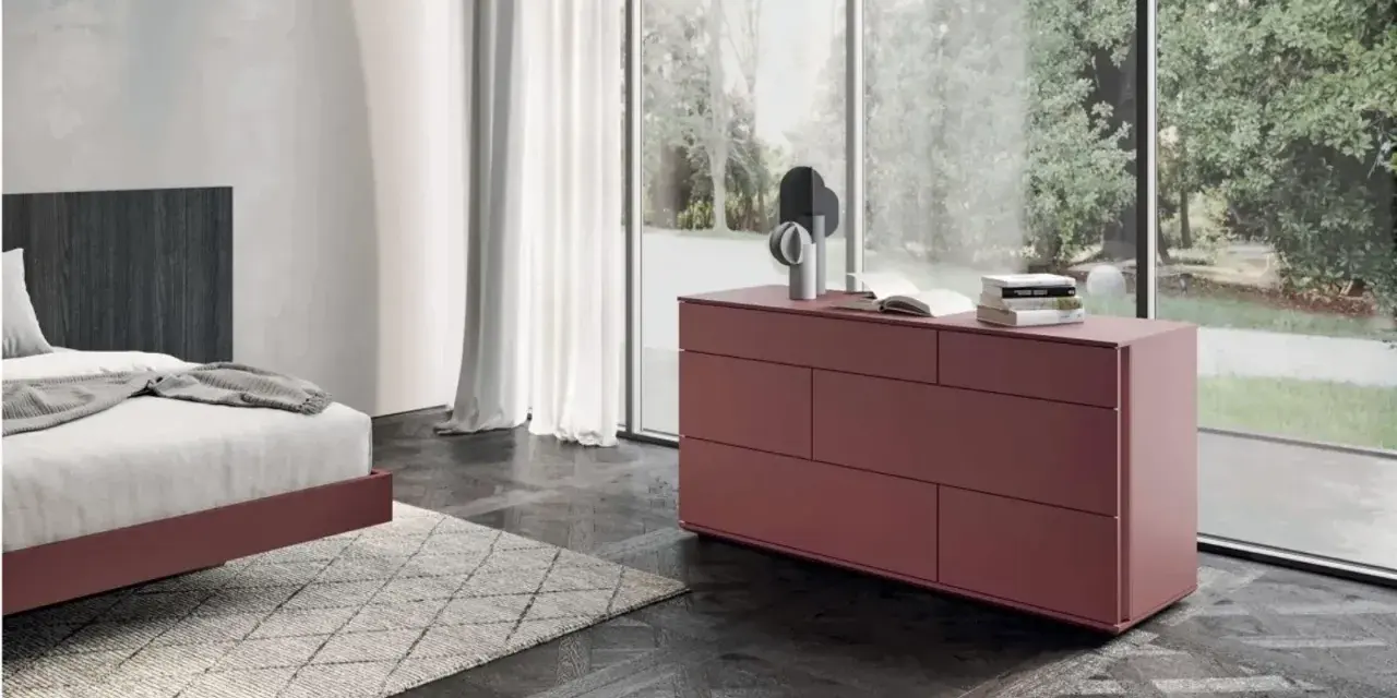 Cassettiera camera da letto moderna color bordeaux, con sei cassetti, accanto a un letto e una grande finestra.