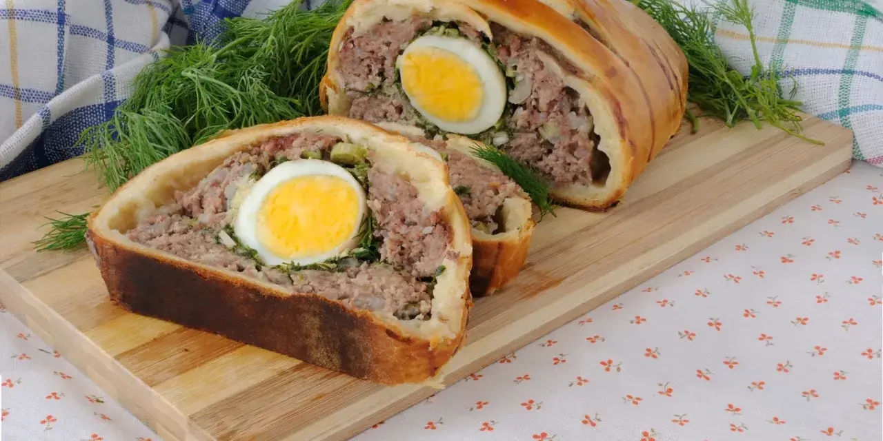 Une tranche de pâté de Pâques, garnie d'un œuf dur, est présentée sur une planche en bois, avec de l'aneth frais.