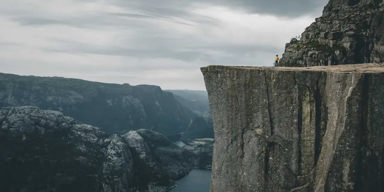 Człowiek w żółtej kurtce stoi na krawędzi skalnego klifu w Norwegii, podziwiając majestatyczny fiord i góry.