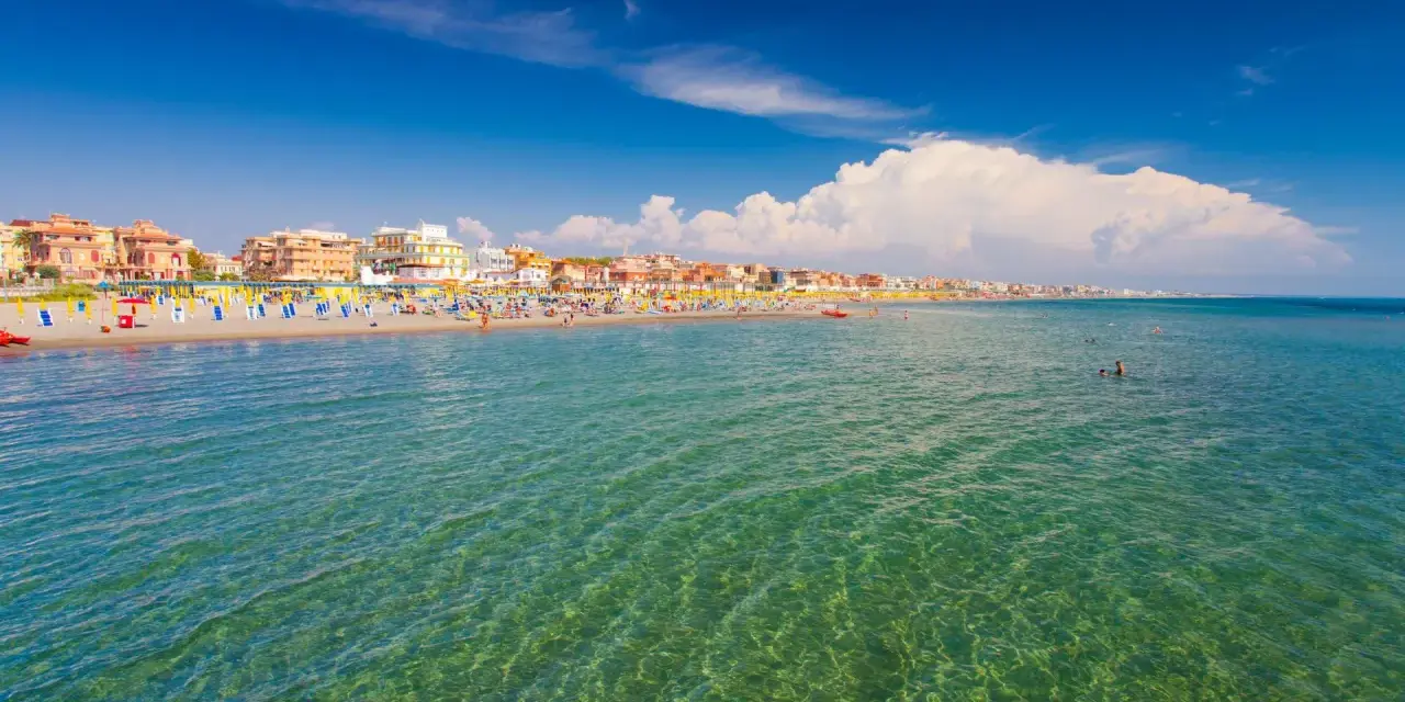 Słoneczna plaża w Rzymie, pełna kolorowych parasoli i ludzi kąpiących się w krystalicznie czystej wodzie.