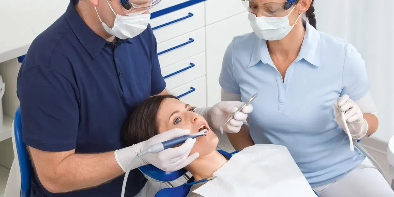 Dentysta i asystentka w okularach i maseczkach pracują przy pacjentce. Asystentka trzyma narzędzie, a dentysta używa **vector paro** do zabiegu.