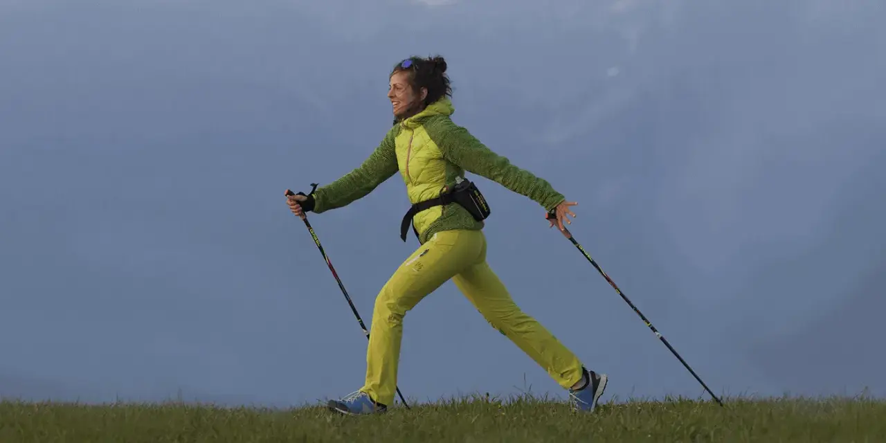 Donna sorridente pratica nordic walking su un prato. Informarsi sulle controindicazioni prima di iniziare.