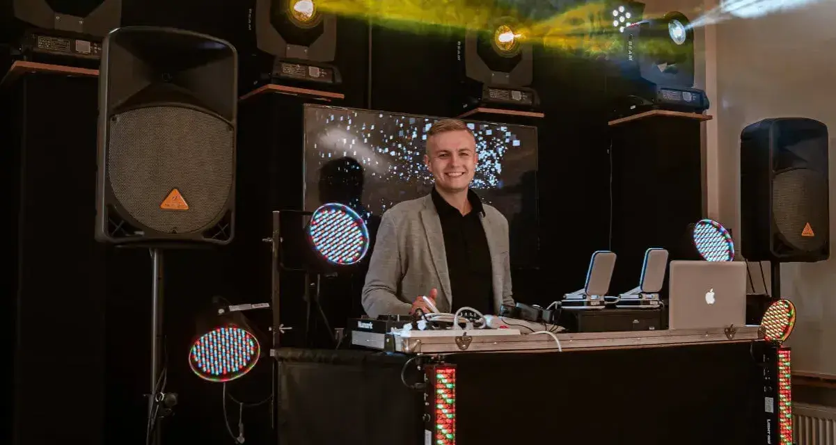 Uśmiechnięty dj na 18. urodziny, gotowy do rozkręcenia imprezy. W tle kolorowe światła i sprzęt nagłośnieniowy.