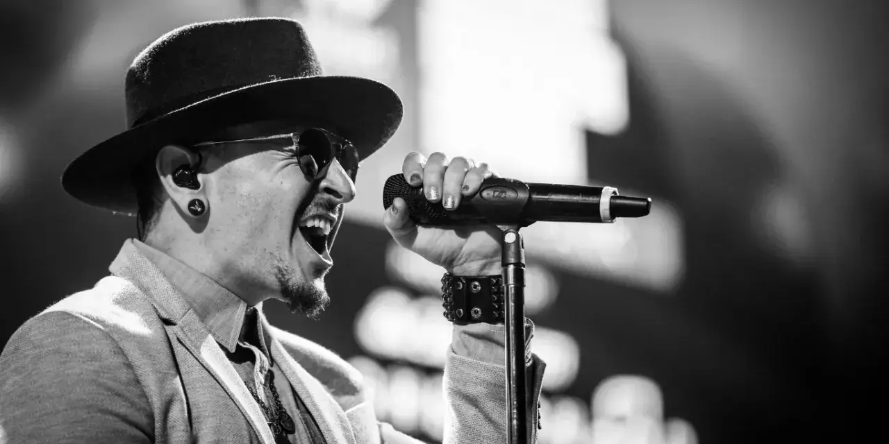 Wokalista Linkin Park, Chester Bennington, w kapeluszu i okularach przeciwsłonecznych, śpiewa do mikrofonu z pasją.